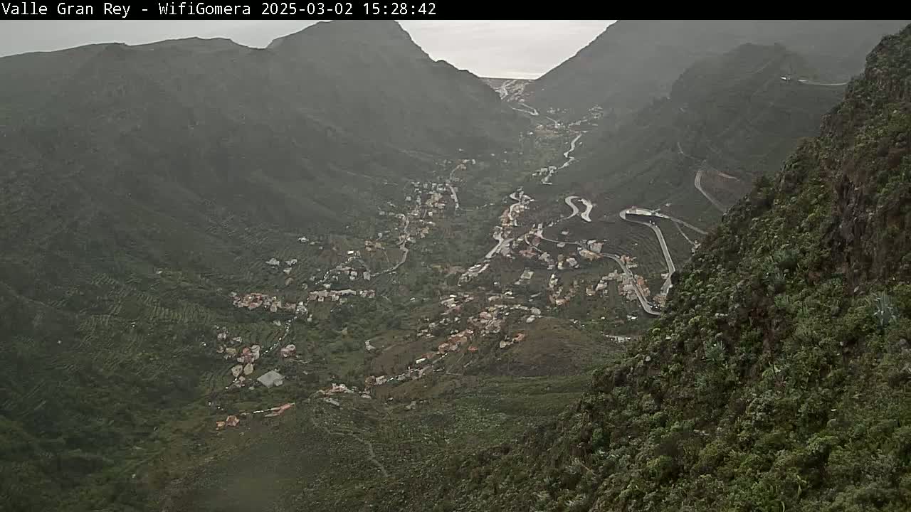 A high-angle view shows a valley town nestled between steep, verdant mountains on an overcast day.