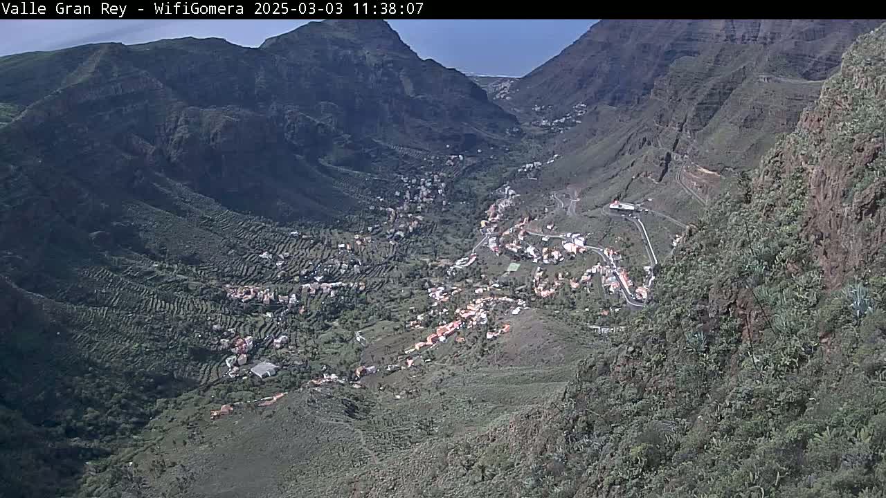 A high-angle view on a sunny day shows a village nestled in a steep, mountainous valley with terraced farmland.
