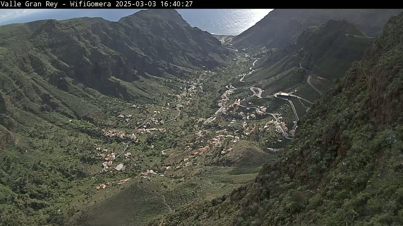 A high-angle view shows a sunny valley nestled between steep, green mountains, with a small town visible in the valley below and a winding road snaking through it.