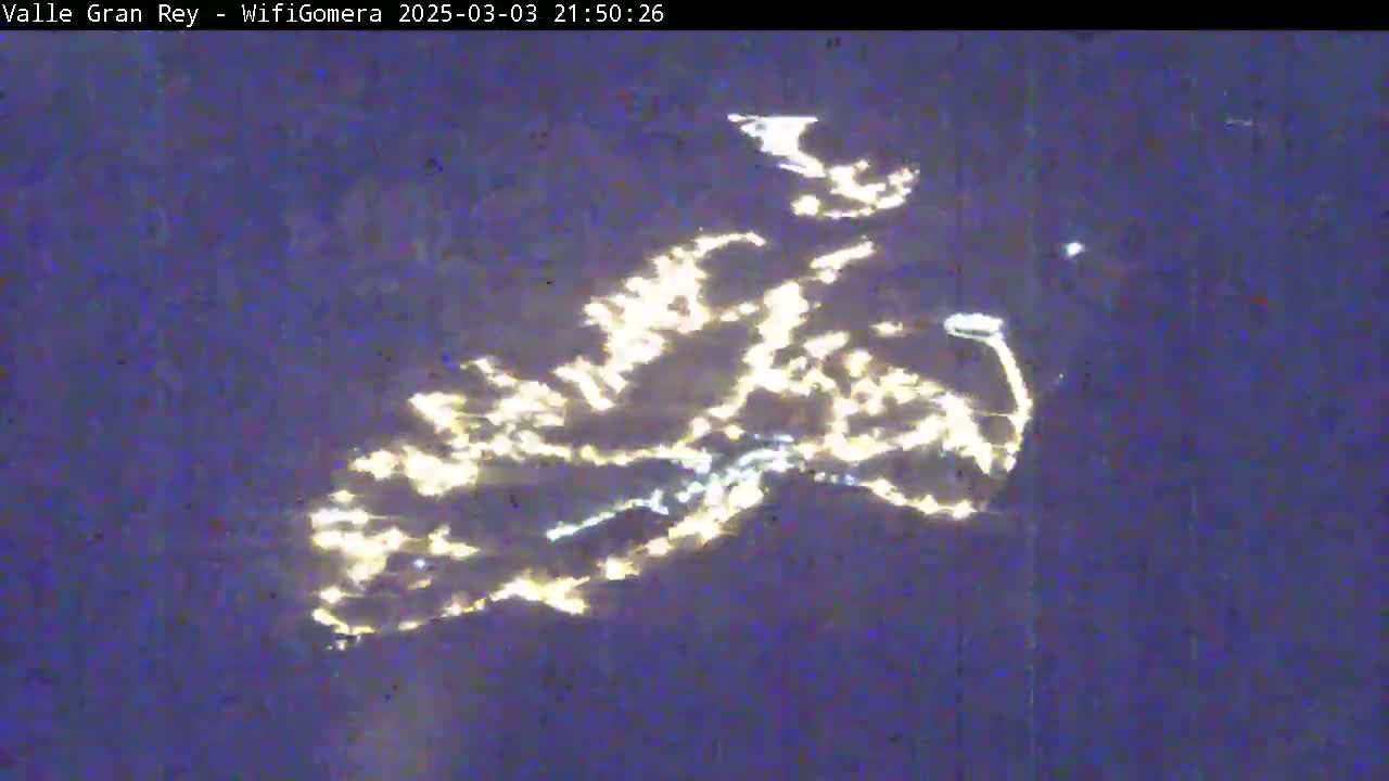 A nighttime aerial view shows the lights of a town or city scattered across a dark, possibly water-covered, landscape.