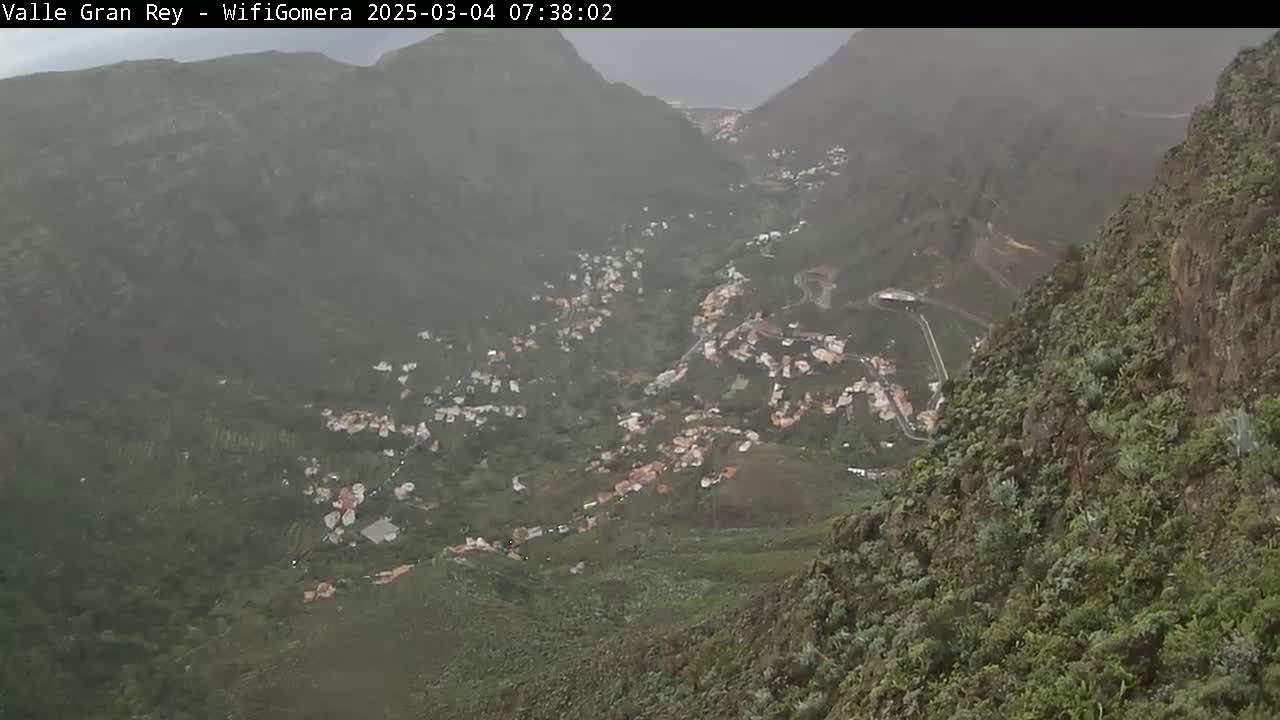 A hazy view shows a town nestled in a verdant valley between steep, mountainous slopes.