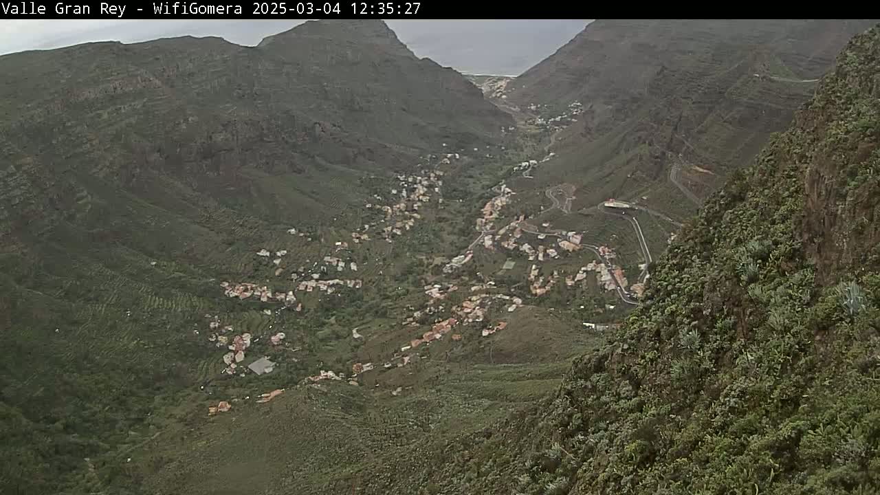 A mountainous valley town is visible under an overcast sky, nestled between steep, verdant hillsides.