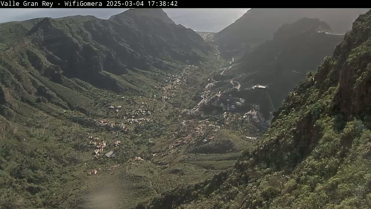 A high-angle view shows a valley nestled between steep, green mountains on a partly sunny day.
