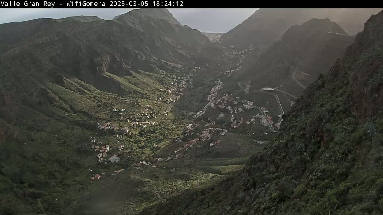 A verdant valley nestled between steep, dark mountains under a hazy sky.