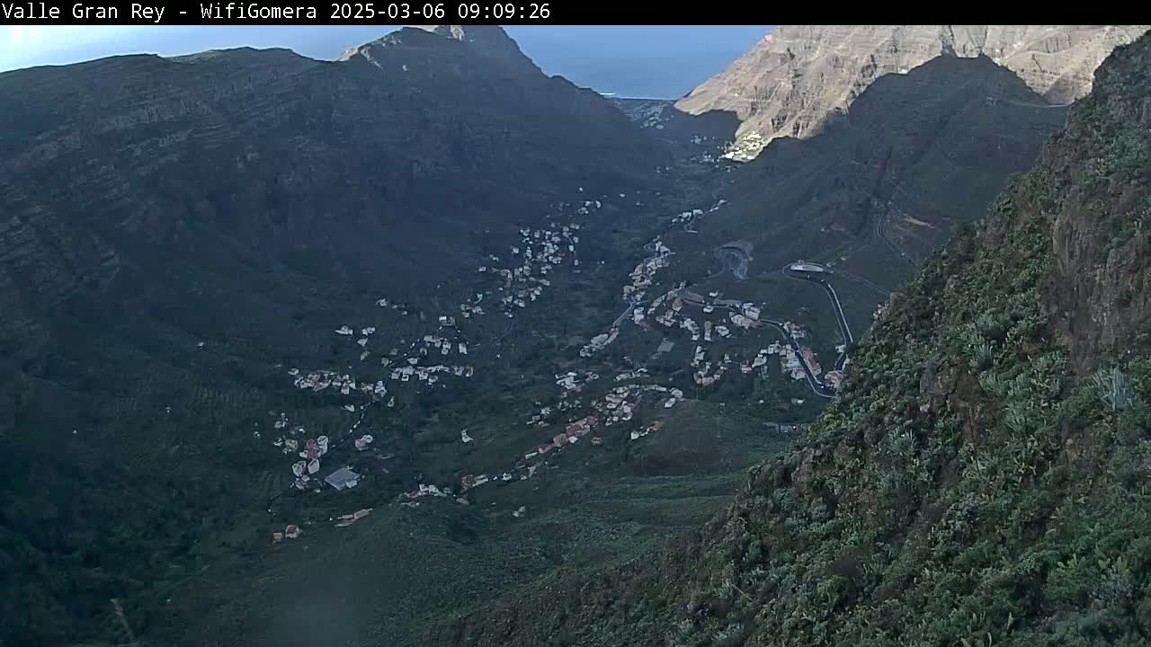 A high-angle view shows a mountainous valley with a village nestled within, under a mostly clear sky.