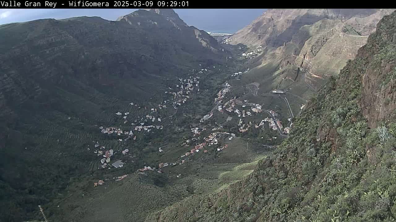 A mountainous valley town is visible under mostly clear skies, nestled between steep, green hillsides.
