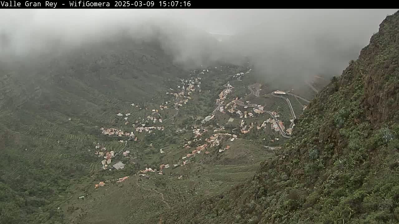A foggy, mountainous valley reveals a village nestled among green hillsides and winding roads.