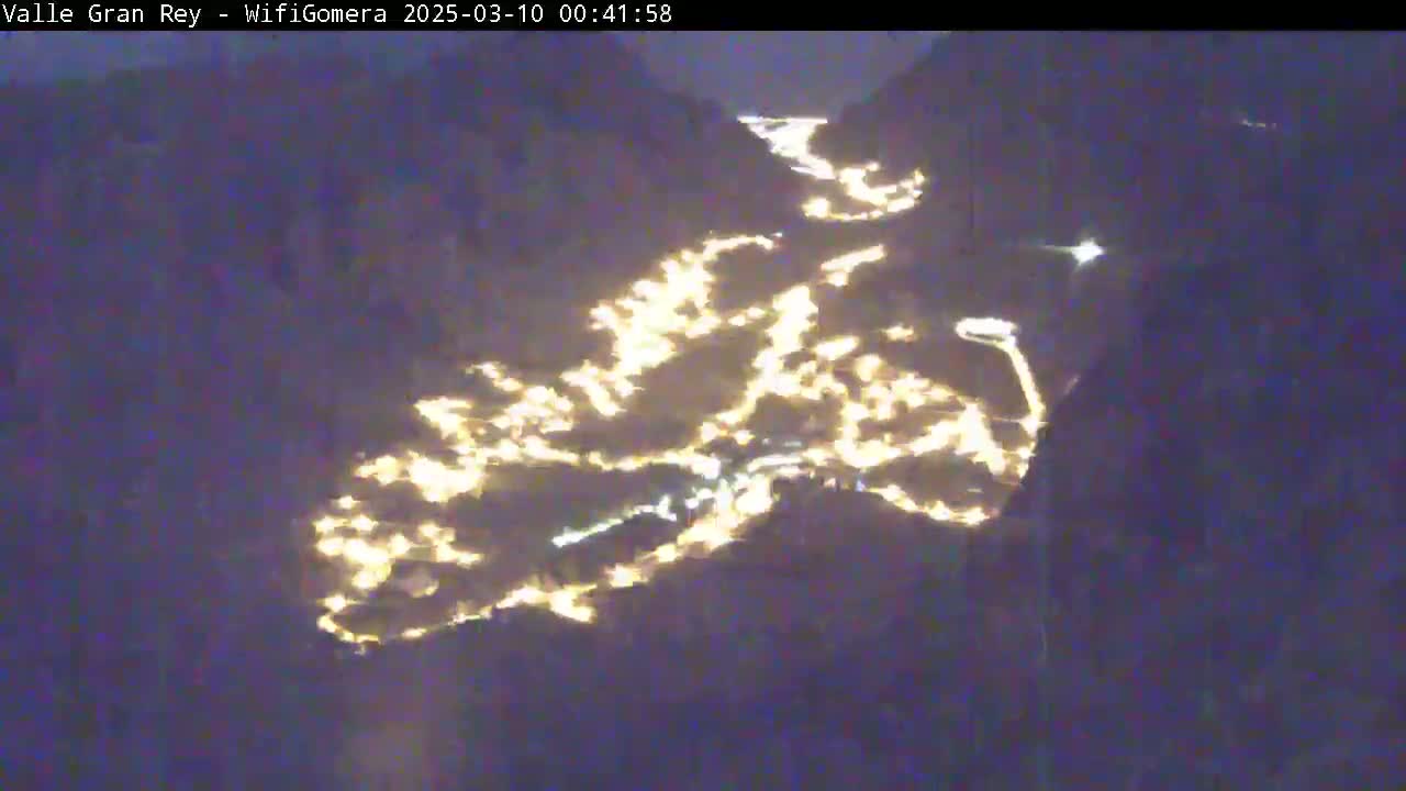 A nighttime, aerial view shows a valley town illuminated by numerous scattered lights nestled between dark mountains.