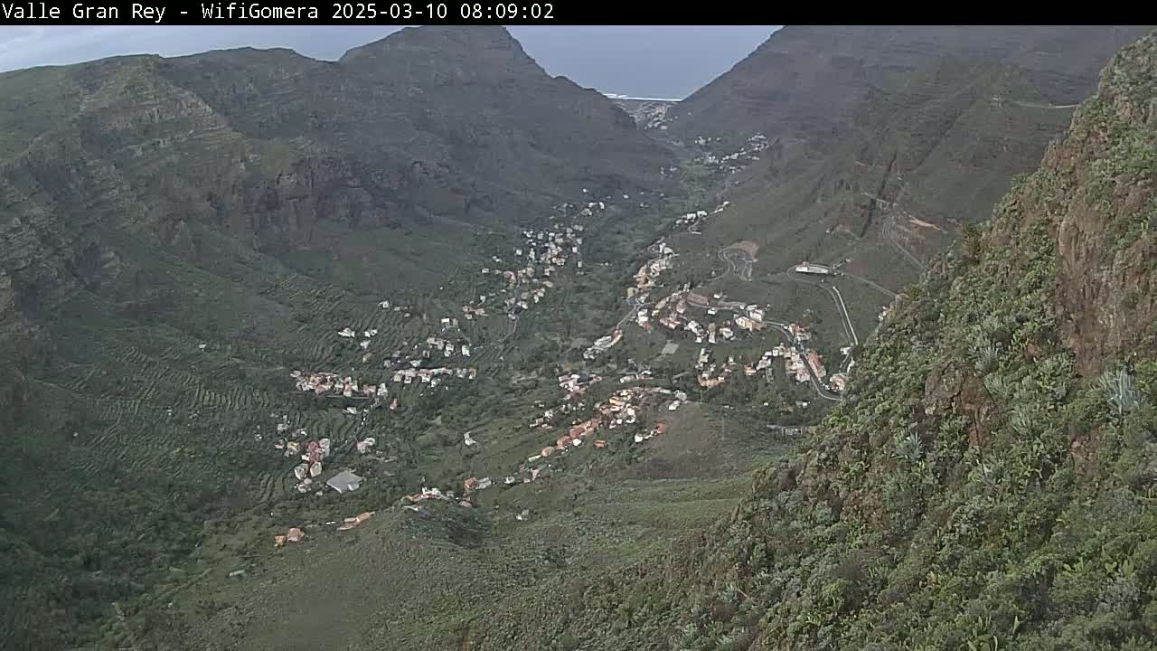 A high-angle view shows a valley town nestled between steep, verdant mountains under a mostly cloudy sky.