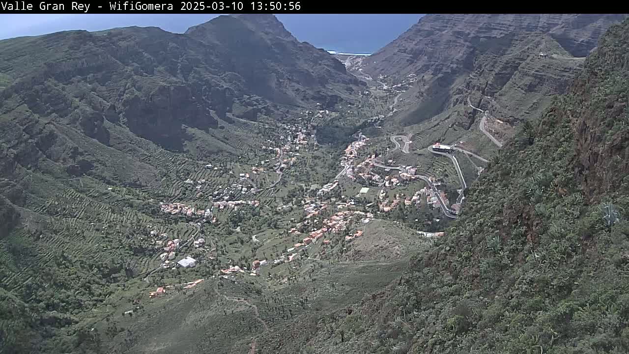 A sunny, high-angle view shows a valley town nestled between steep, mountainous terrain, with winding roads snaking through the landscape.