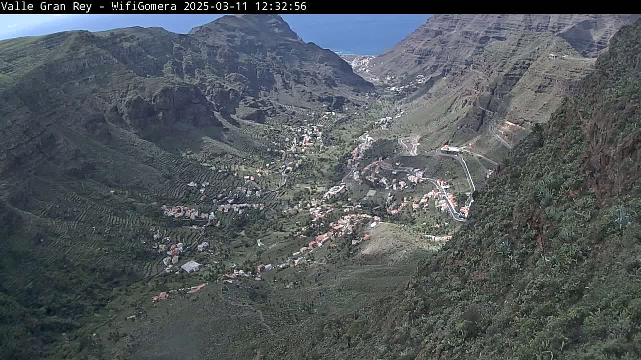 A high-angle view shows a valley town nestled amongst steep, terraced hillsides on a sunny day, with a glimpse of ocean in the far distance.