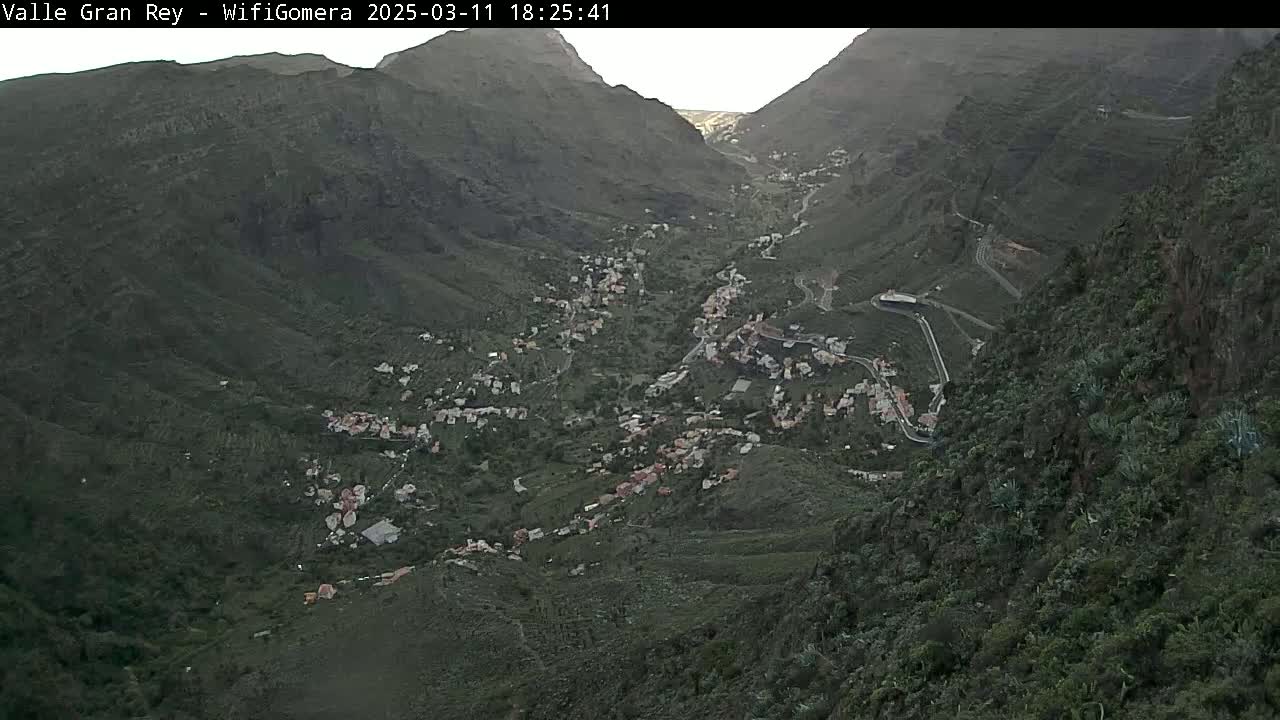 A high-angle, long shot shows a town nestled in a valley between dark, mountainous terrain under overcast skies.