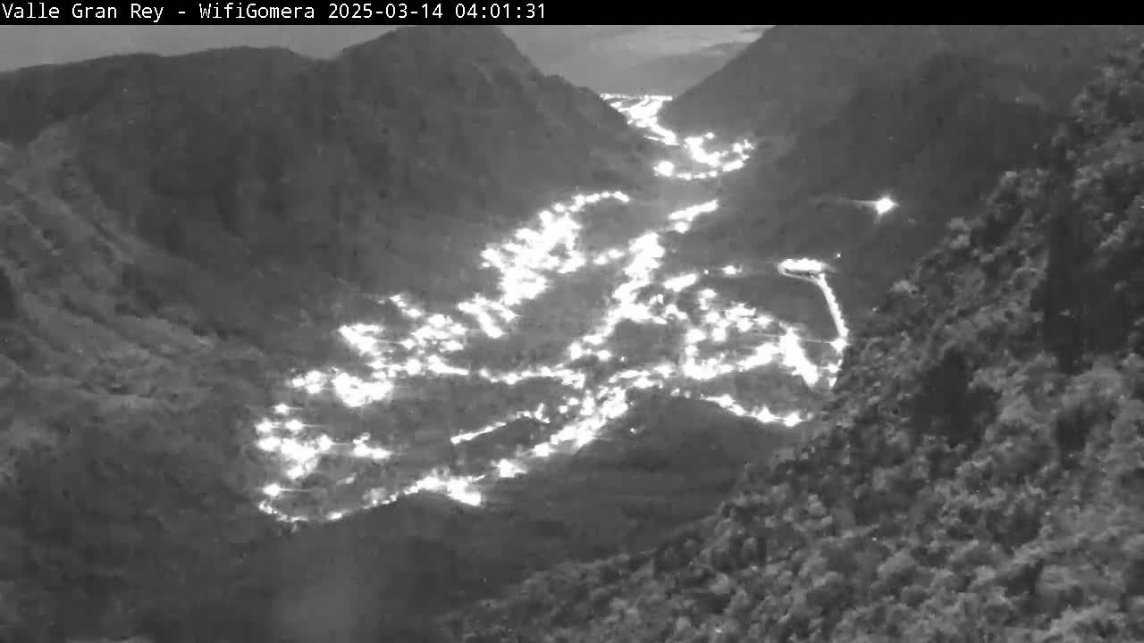 A nighttime, high-angle view shows a valley with many lights from a town nestled between dark mountains.