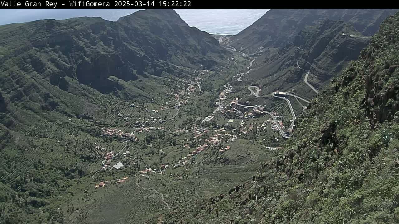 A high-angle view shows a valley town nestled between steep, green mountains, with a glimpse of the ocean in the distance under sunny conditions.