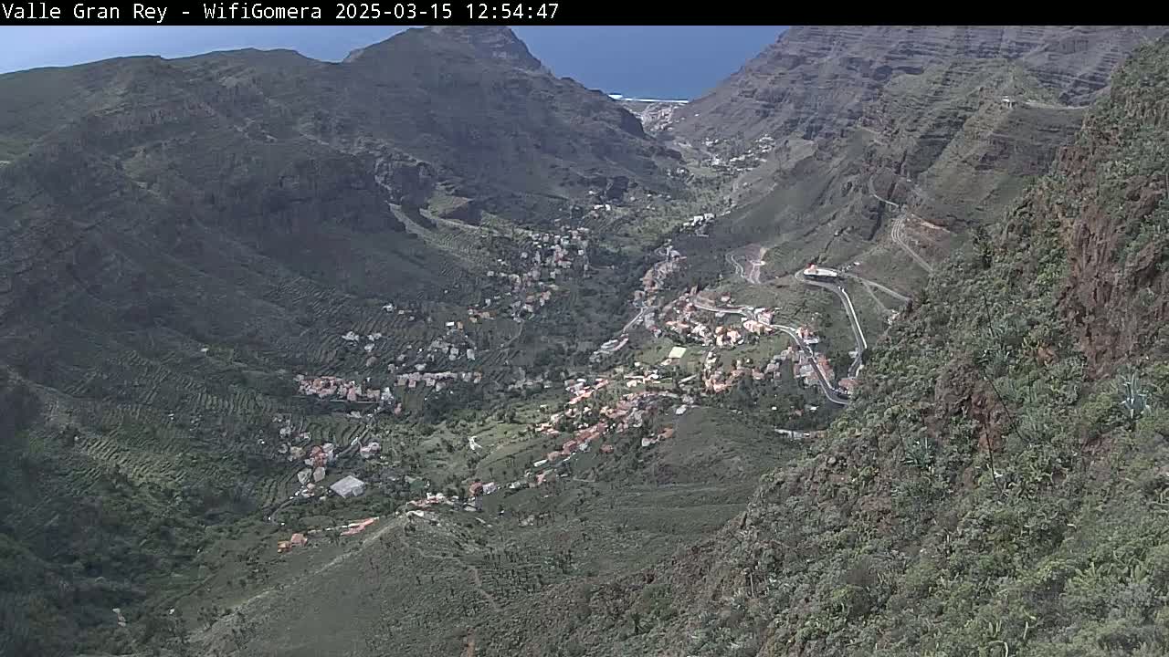 A high-angle, long shot shows a village nestled in a mountainous valley on a sunny day.