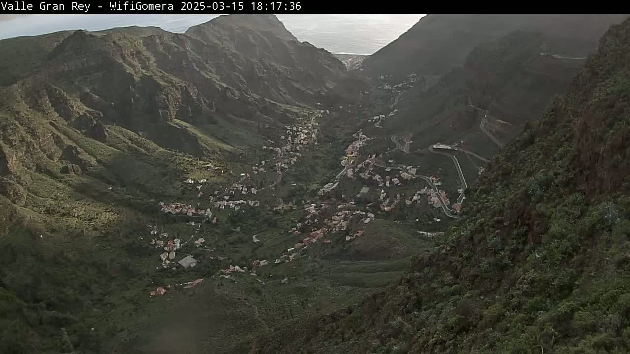 A high-angle view shows a town nestled in a verdant valley between steep, rocky mountainsides under a partly cloudy sky.