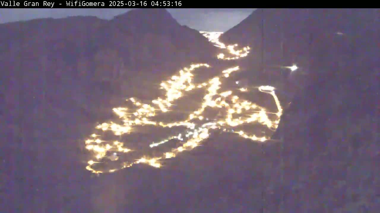 A nighttime aerial view shows a valley town's lights winding along a road between dark mountains.