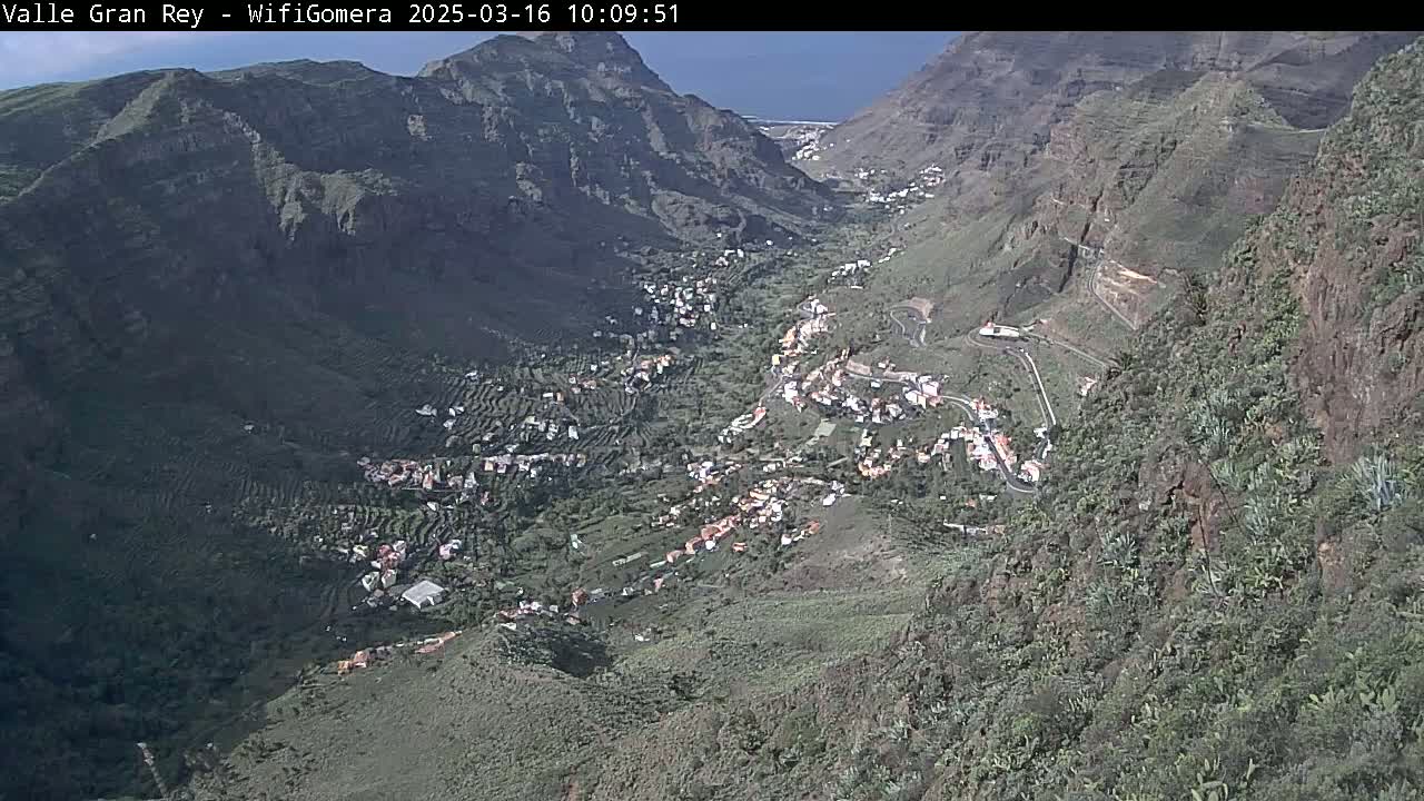 A sunny, high-angle view shows a valley town nestled between steep, rocky mountainsides, with vegetation covering the slopes and some roads winding through the landscape.