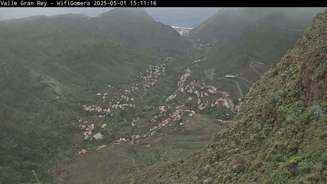 A hazy view from a mountaintop shows a village nestled in a verdant valley between steep, rocky slopes.