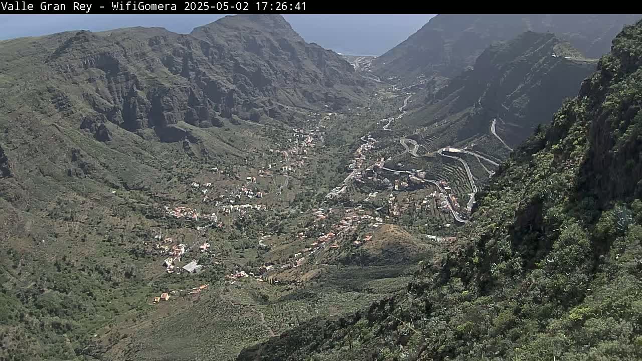 A sunny, high-angle view shows a village nestled in a verdant valley between steep, rocky mountainsides.