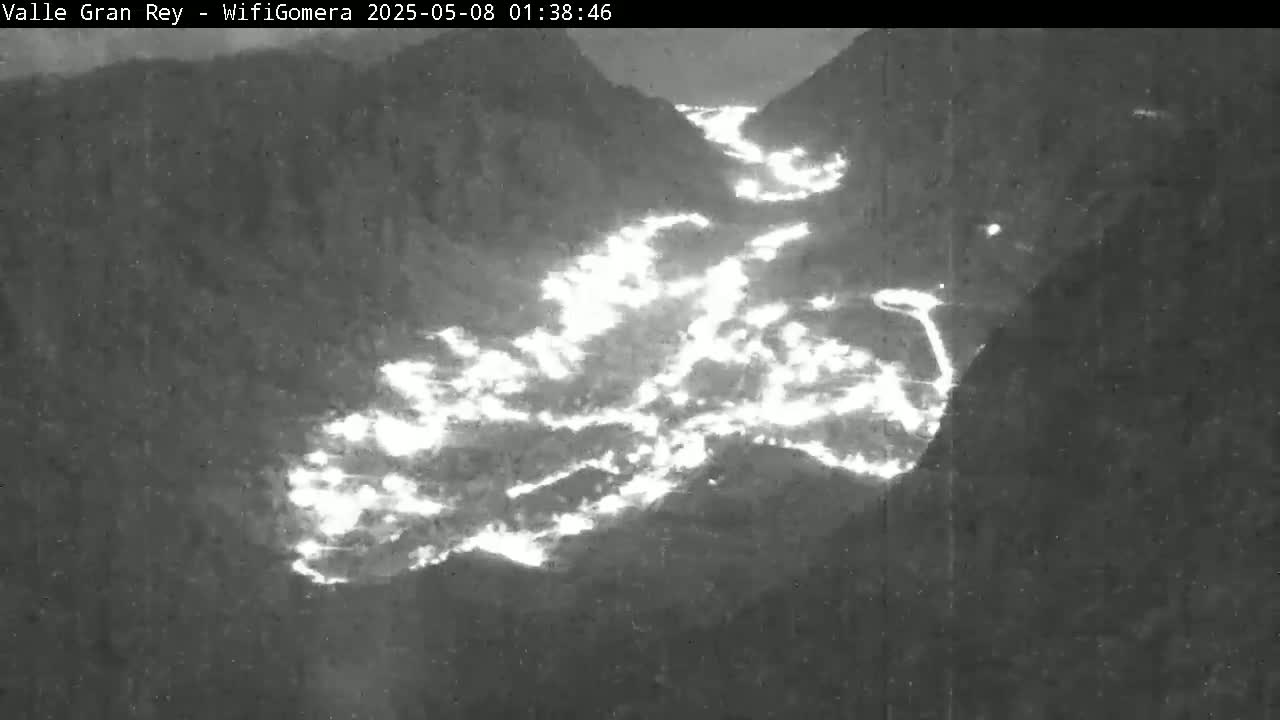 A nighttime, high-angle view shows a valley illuminated by bright, sporadic lights flowing down a mountainous region.