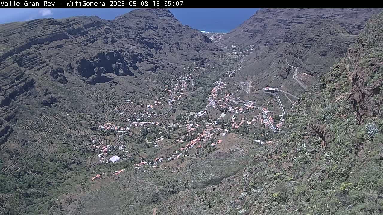 A high-angle, sunny view shows a small town nestled in a valley between steep, rocky mountains.