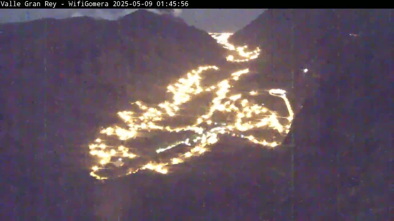 A nighttime, aerial view shows a valley nestled between dark mountains, illuminated by numerous scattered lights.