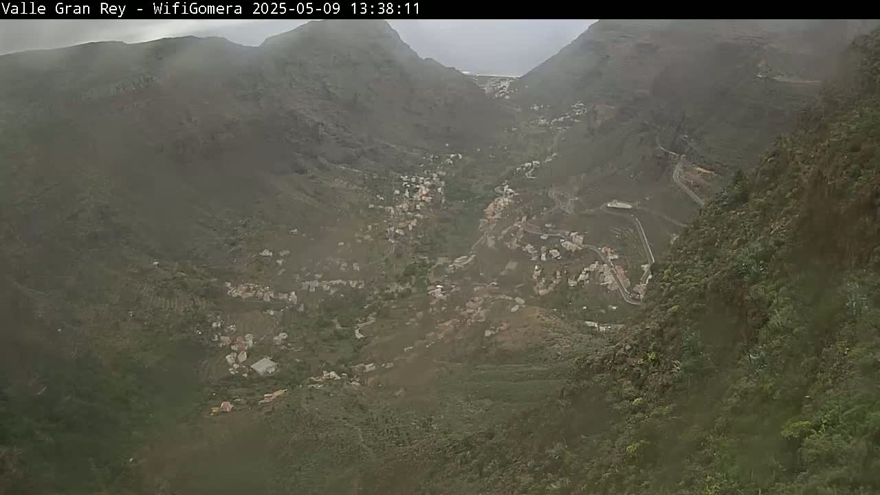 A hazy, overcast view shows a valley nestled between steep, dark green mountains, with a small village visible in the valley floor.
