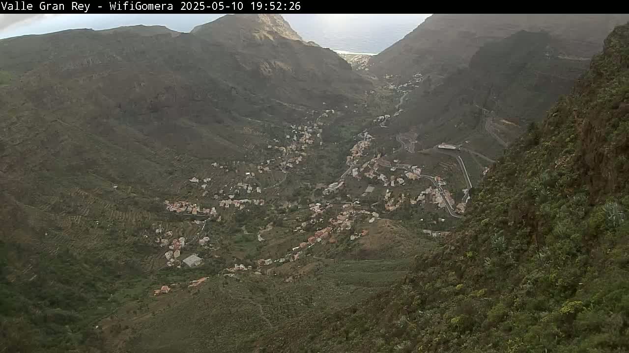 A high-angle view shows a village nestled in a verdant valley between steep, rocky mountains, with a glimpse of the ocean in the distance under hazy skies.