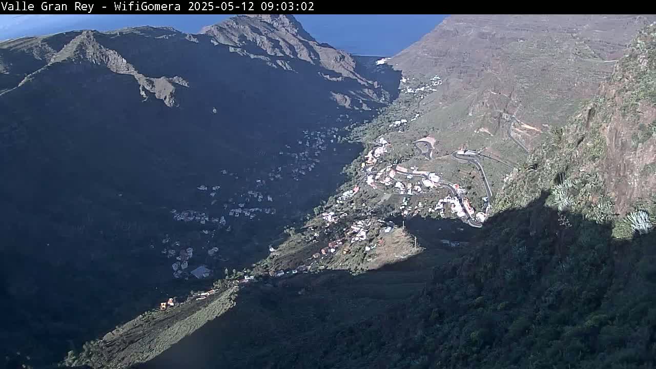 A sunny, high-angle view shows a village nestled in a deep valley between steep, dark mountains.