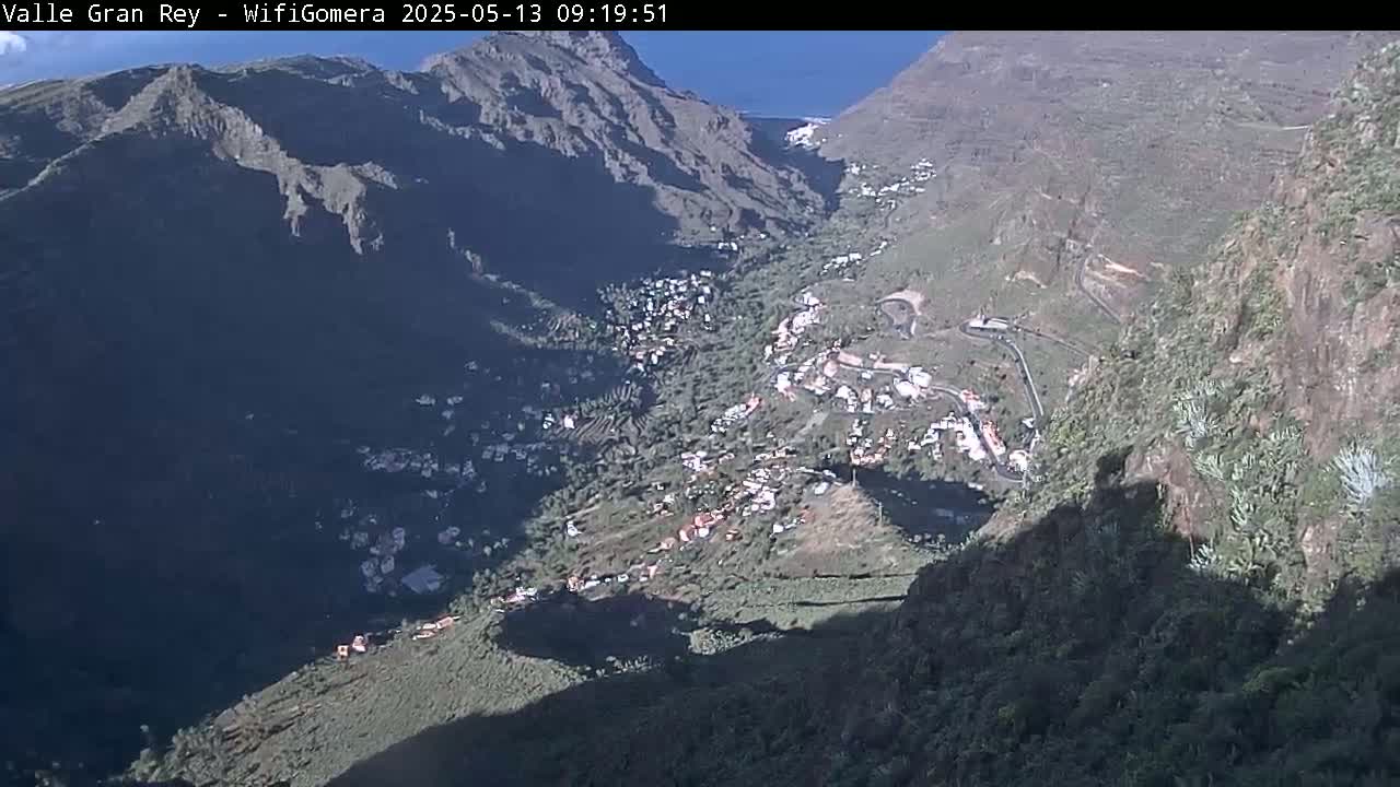 A high-angle, sunny view shows a village nestled in a deep valley between steep, rocky mountains.