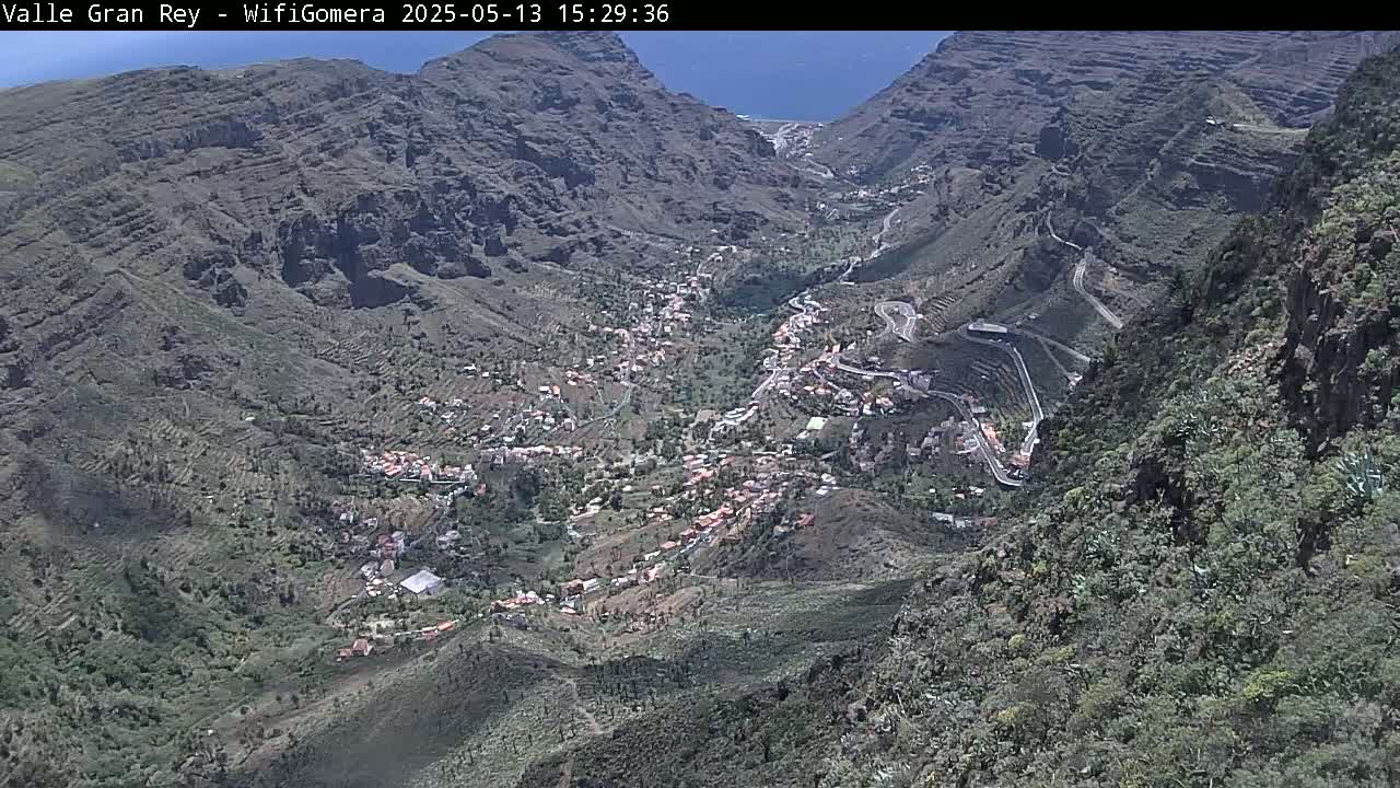 A high-angle view shows a valley town nestled between steep, rocky mountainsides on a clear day.