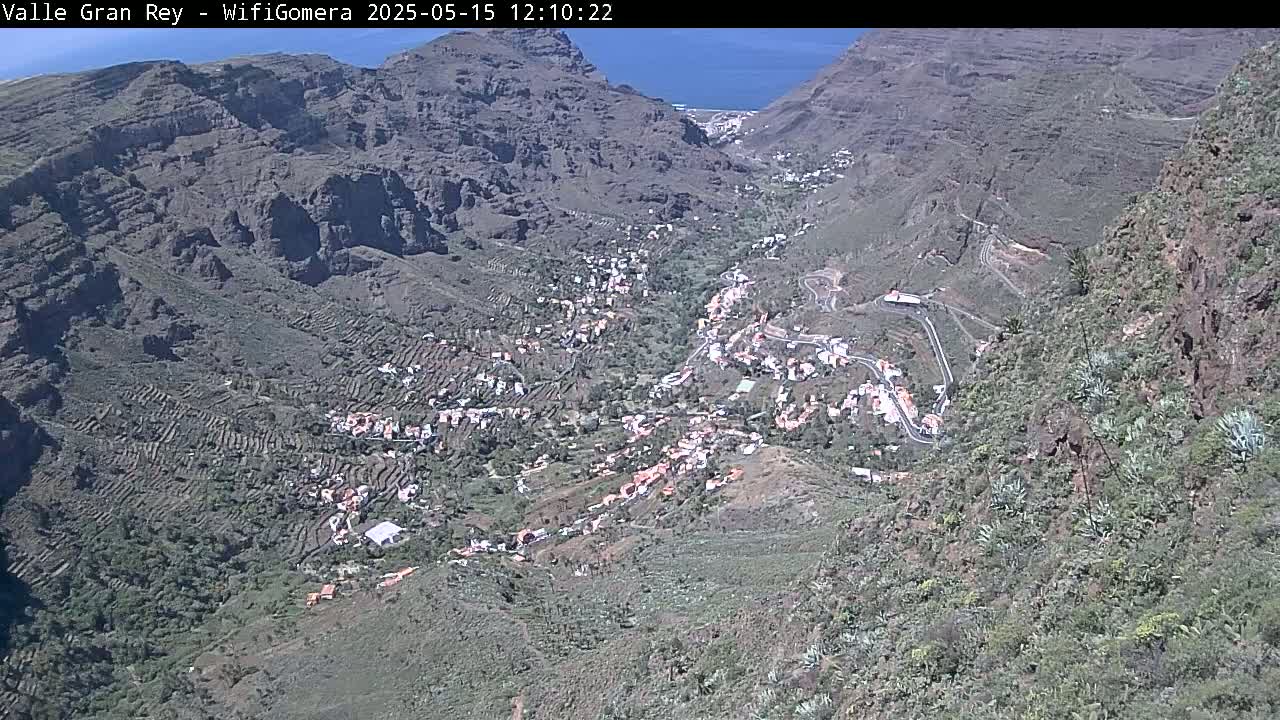 An aerial view shows a village nestled in a mountainous valley on a sunny day, with the ocean visible in the distance.