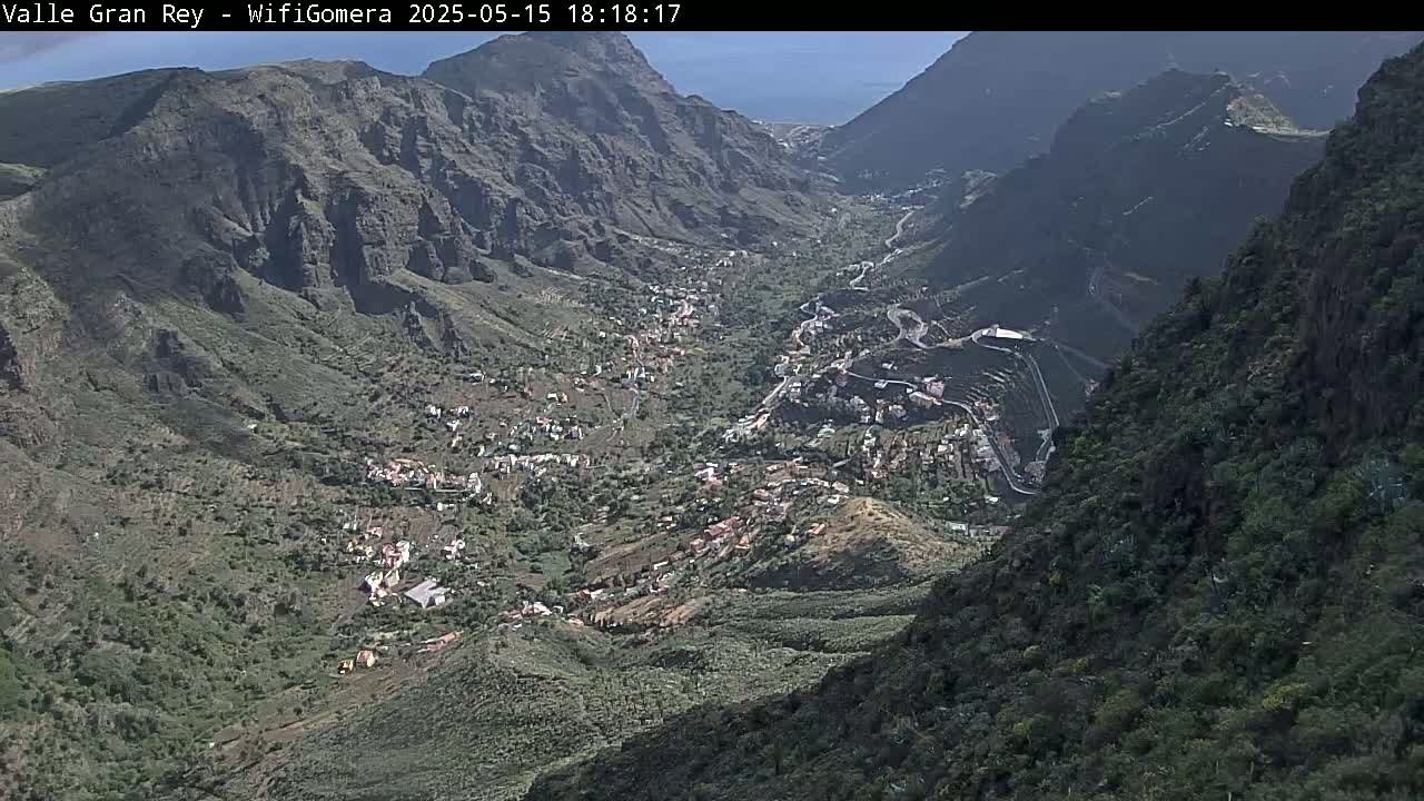 A high-angle view shows a village nestled in a verdant valley between steep, dark-colored mountains on a sunny day.