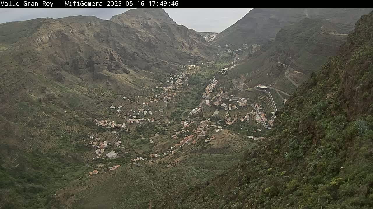 An aerial view shows a town nestled in a mountainous valley under an overcast sky.