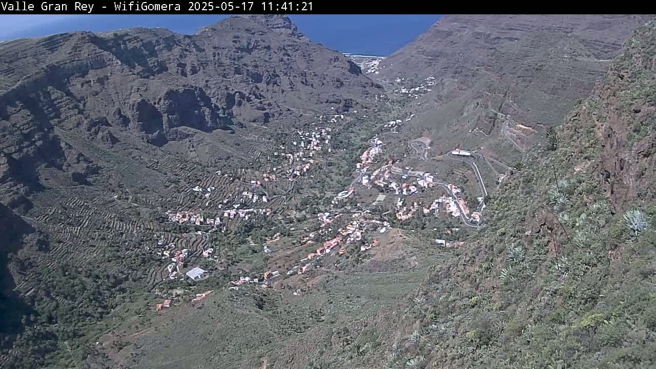 A valley town nestled between steep, rocky mountains on a sunny day is viewed from above.