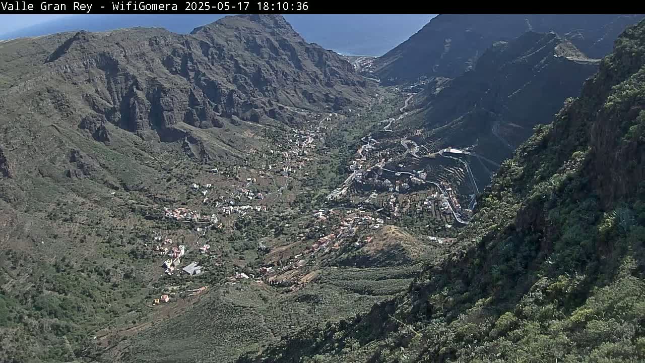 A high-angle view shows a sunny, mountainous valley with a small town nestled within, winding roads snaking through the landscape.