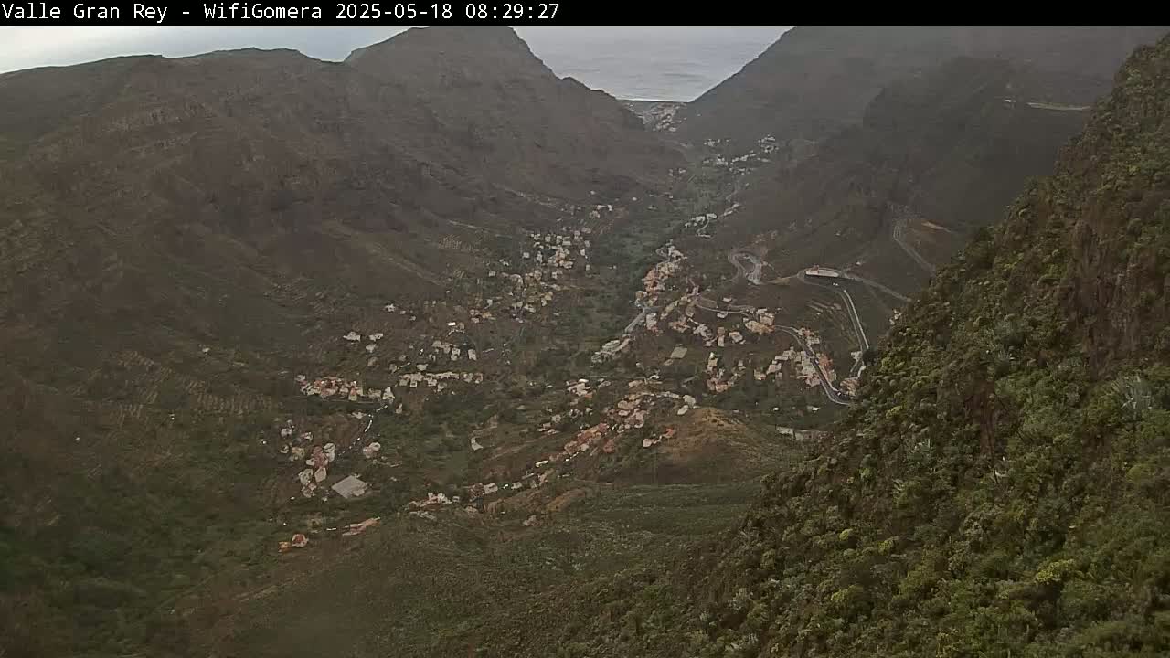 A high-angle view shows a valley town nestled between steep, green mountains under an overcast sky.