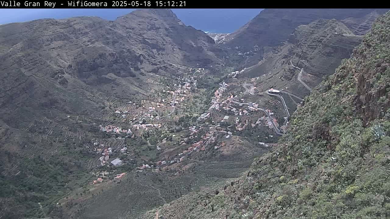 A high-angle view shows a town nestled in a valley between steep, rocky mountains under a mostly clear sky.