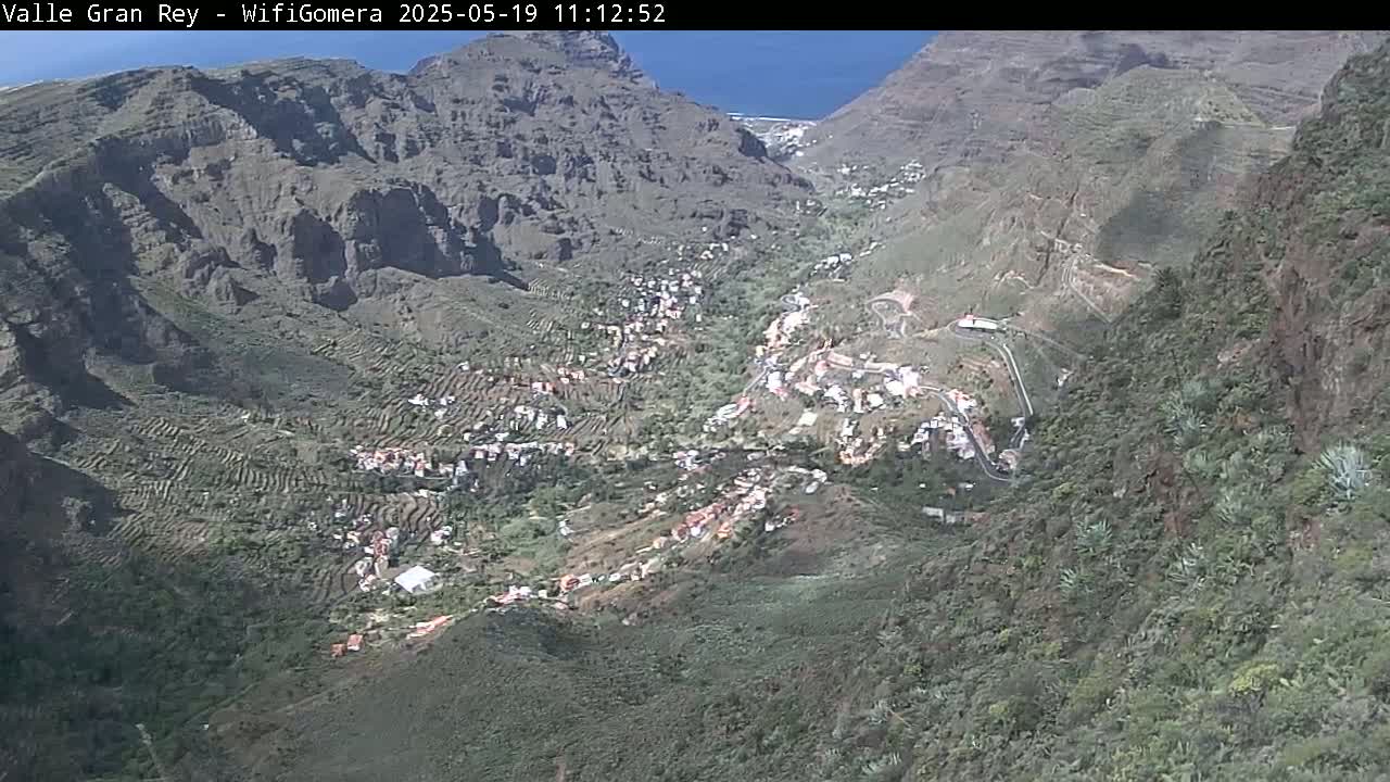 A high-angle view shows a village nestled in a steep, rocky valley on a sunny day, with the ocean visible in the distance.