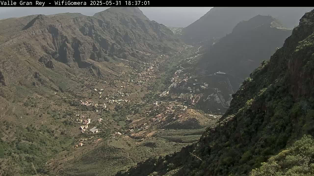 A hazy, high-angle view shows a valley town nestled between steep, rocky mountainsides.