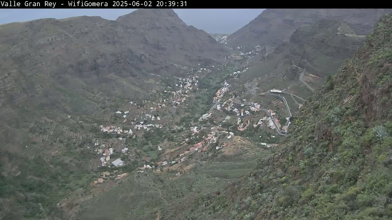 An aerial view shows a village nestled in a valley between steep, rocky mountains under an overcast sky.