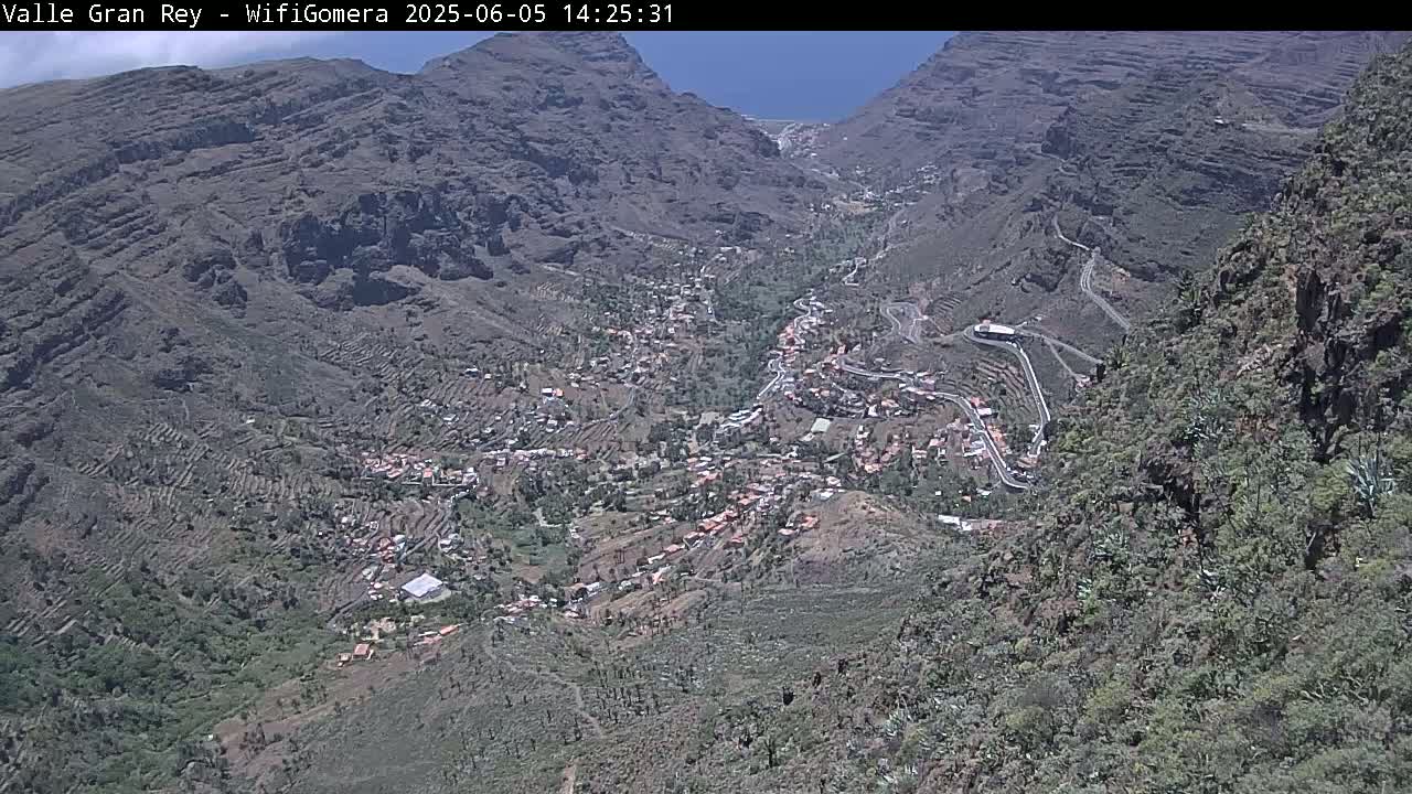 A high-angle, sunny view shows a village nestled in a mountainous valley with terraced farmland.
