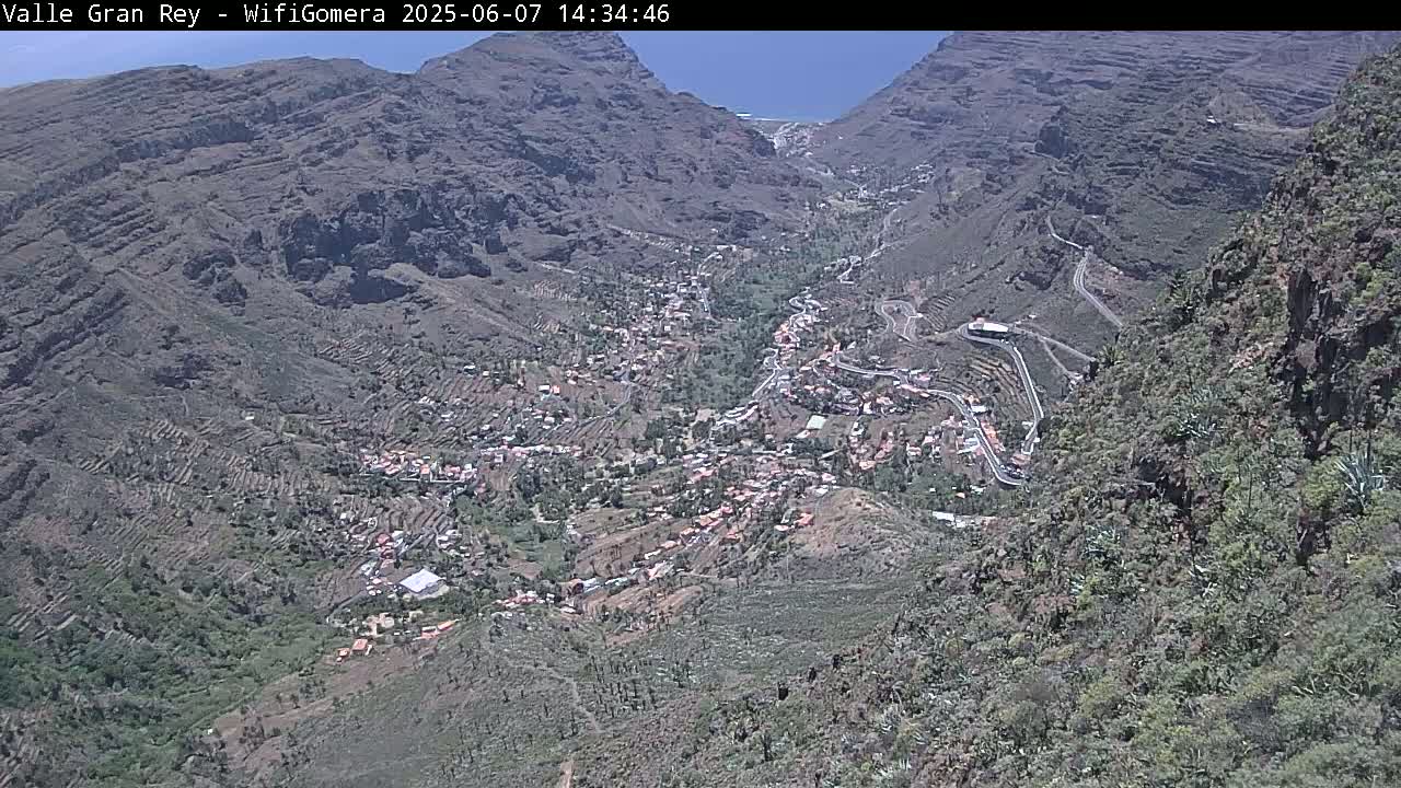 A high-angle view on a sunny day shows a small town nestled in a valley between steep, rocky mountains with winding roads and terraced farmland.