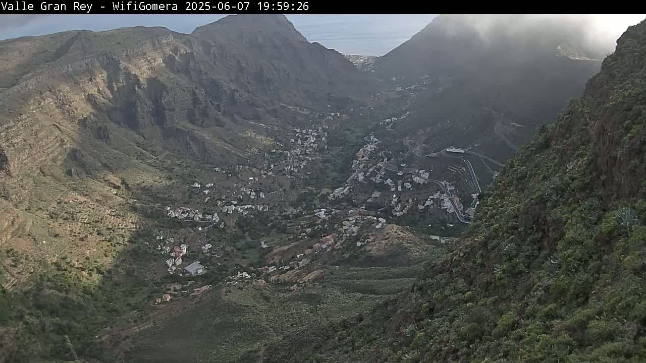 A mountain valley town is nestled between steep, rocky hillsides under partly cloudy skies.