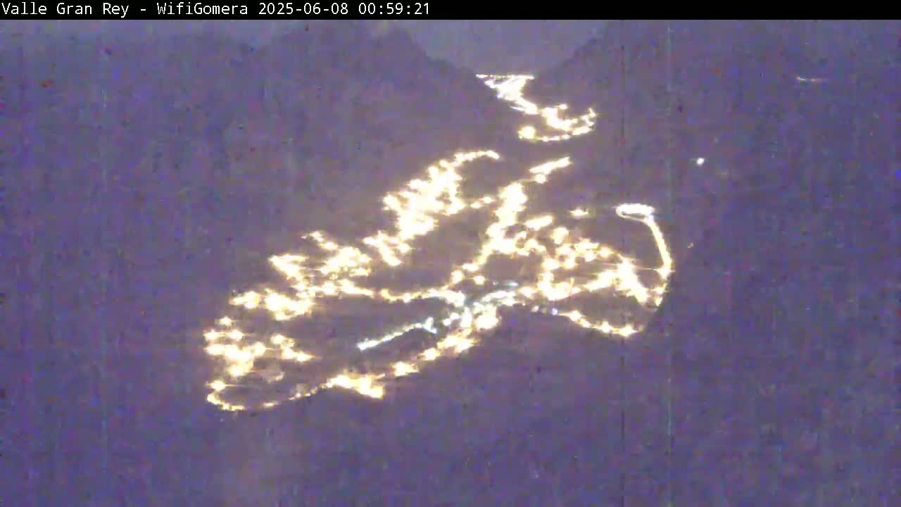 A nighttime aerial view shows a town nestled in a valley, illuminated by numerous lights.