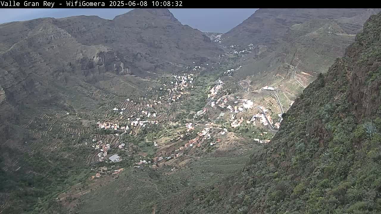 A mountainous valley with terraced farmland and a small village is viewed from above under overcast skies.