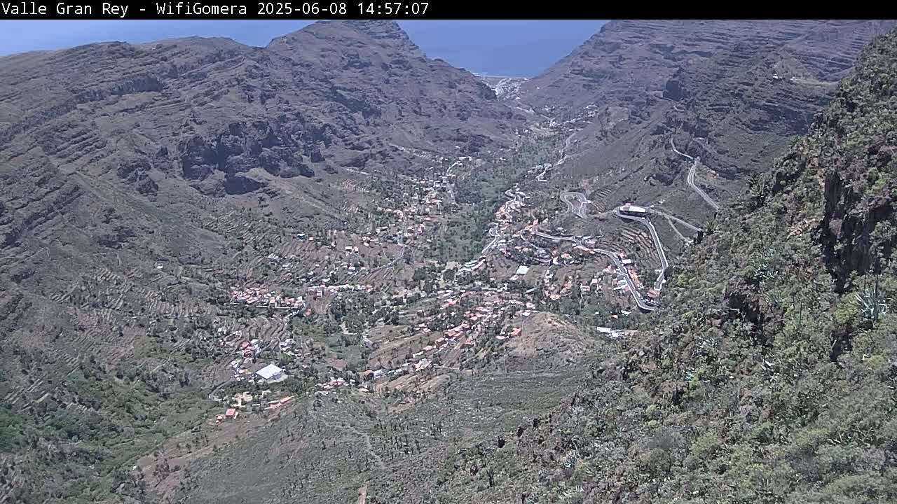 A high-angle view on a sunny day shows a village nestled in a mountainous valley with winding roads snaking through the terrain.