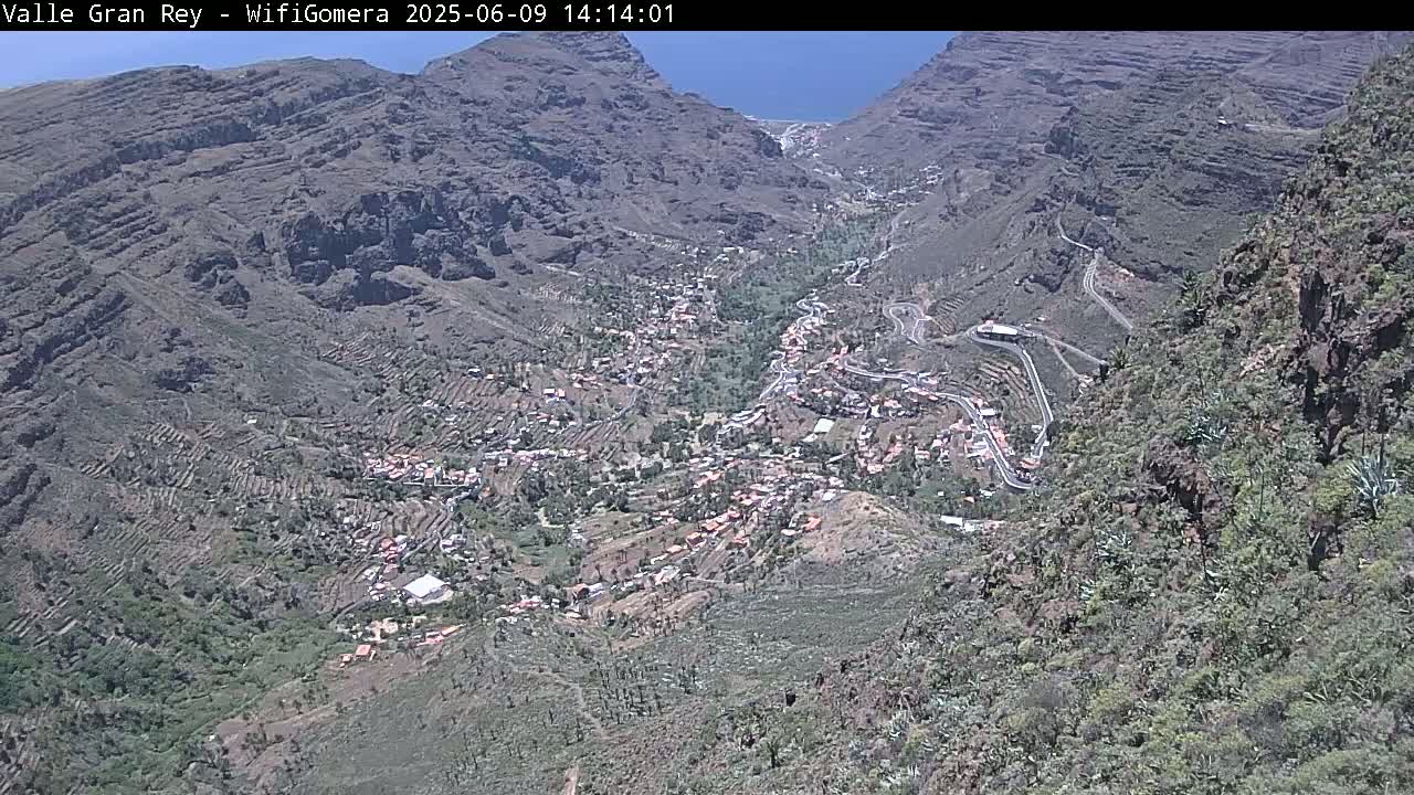 A high-angle view shows a village nestled in a steep, rocky valley on a sunny day.