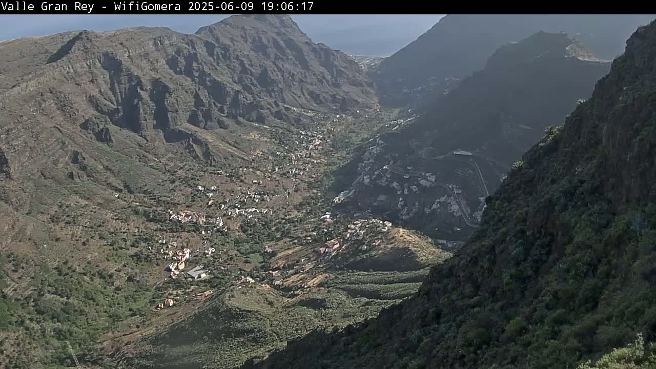 A high-angle view shows a sunlit valley nestled between steep, rocky mountains, with a small village visible in the valley below.